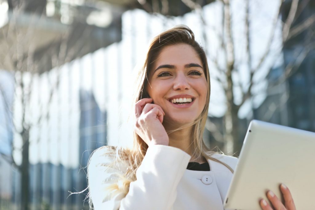 Image of woman in a white top using a mobile phone outside while holding a tablet