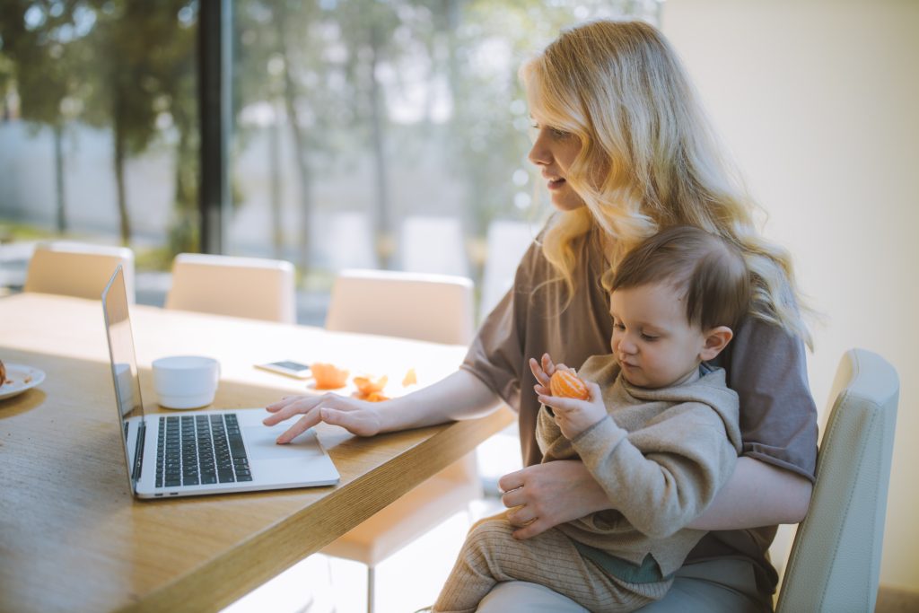 Woman sat at kitchen table with a silver laptop holding a baby eating an orange in one arm