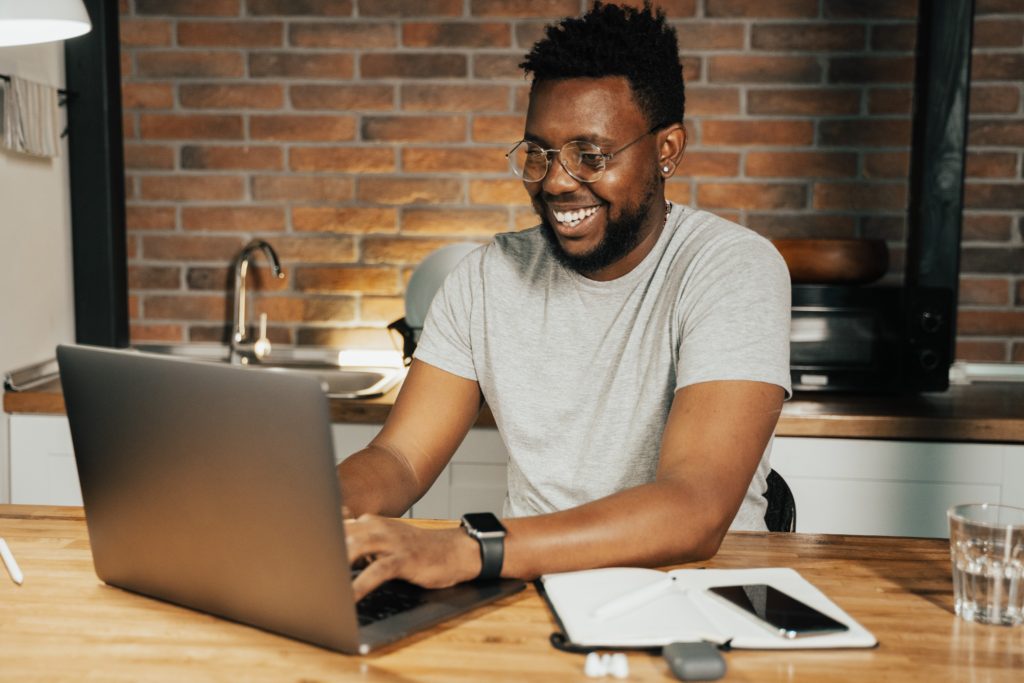 A smiling man sat at wooden table in kitchen working on a silver laptop woth a notepad and black phone next to him