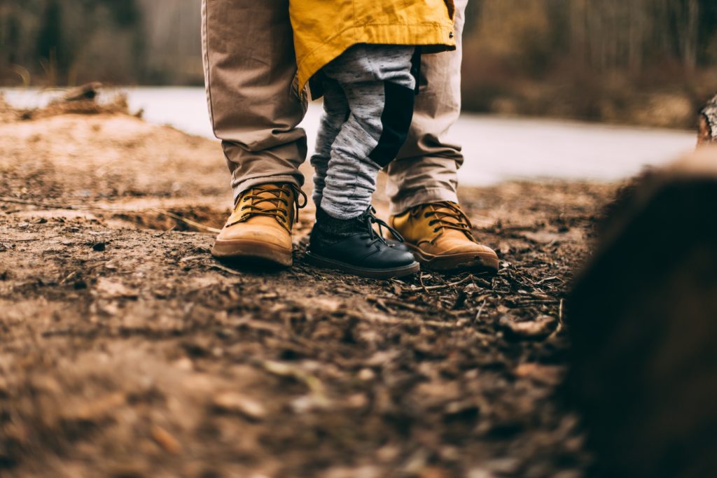 Image shows an adult's feet wearing brown walking shoes with a child next to them wearing black shoes and a yellow coat