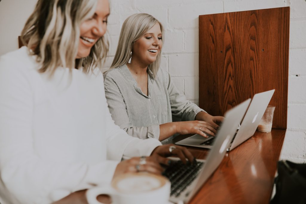 Two women with blonde hair, smiling, sat working at silver laptops on a brown wooden desk