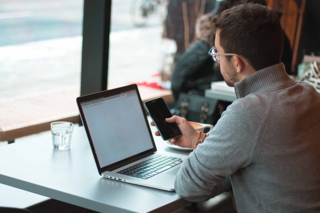 Man sat at a table in a coffee shop with a phone for work infront of him and a silver laptop on the grey table he is sat at