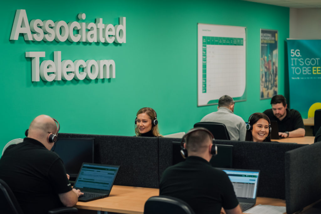 The Associated Telecom team sitting at light wood desks , laptops in front of them, black headsets on and a green wall behind them with the words Associated Telecom behind them.