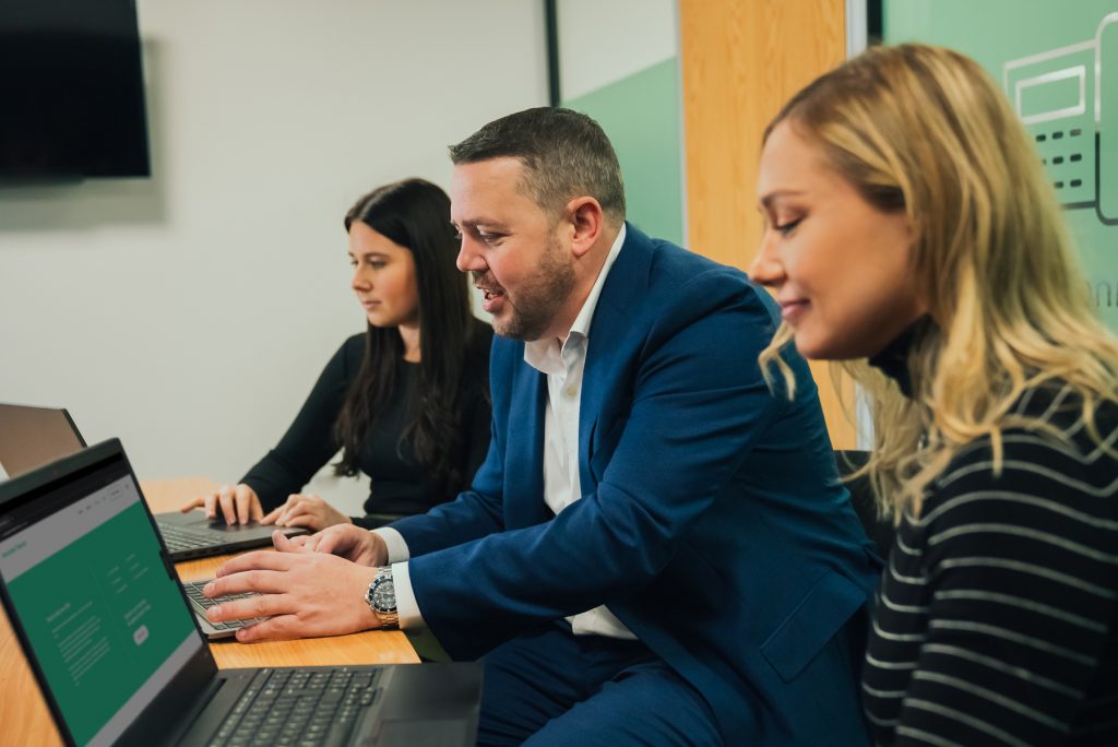 image of 3 members of staff at Associated Telecom sat at a wooden desk using black laptops