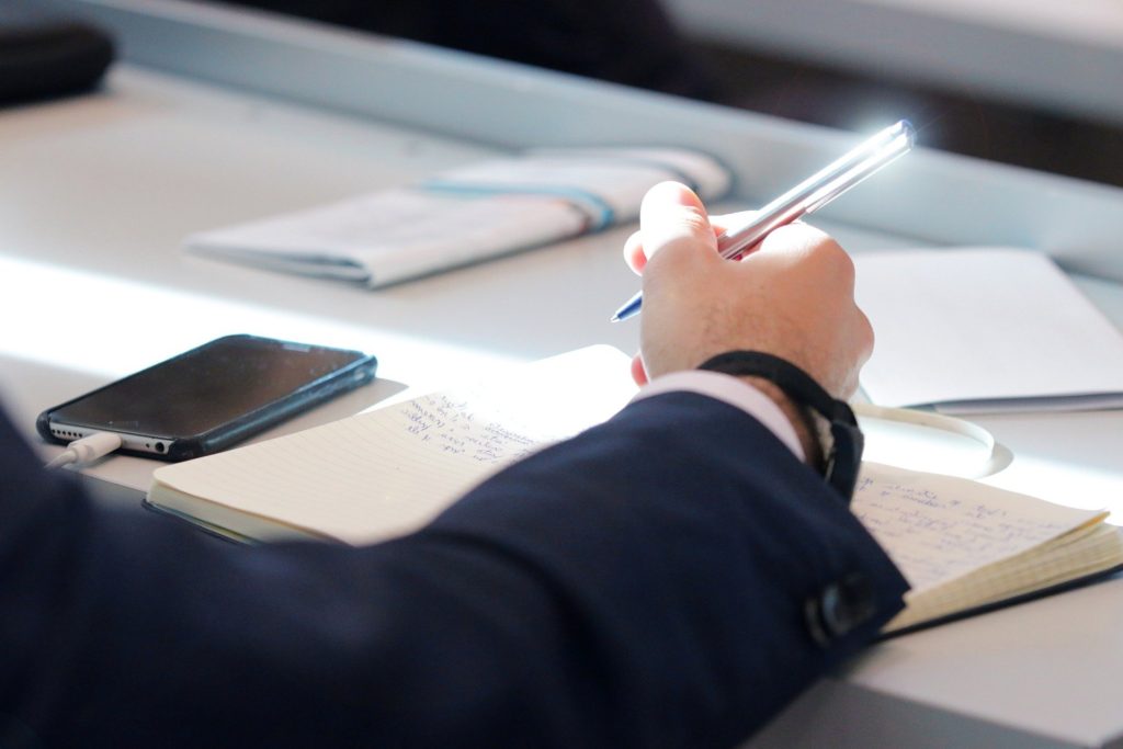 Image features a man in smart clothing, taking a break from writing in his notebook, looking at his nearby phone.