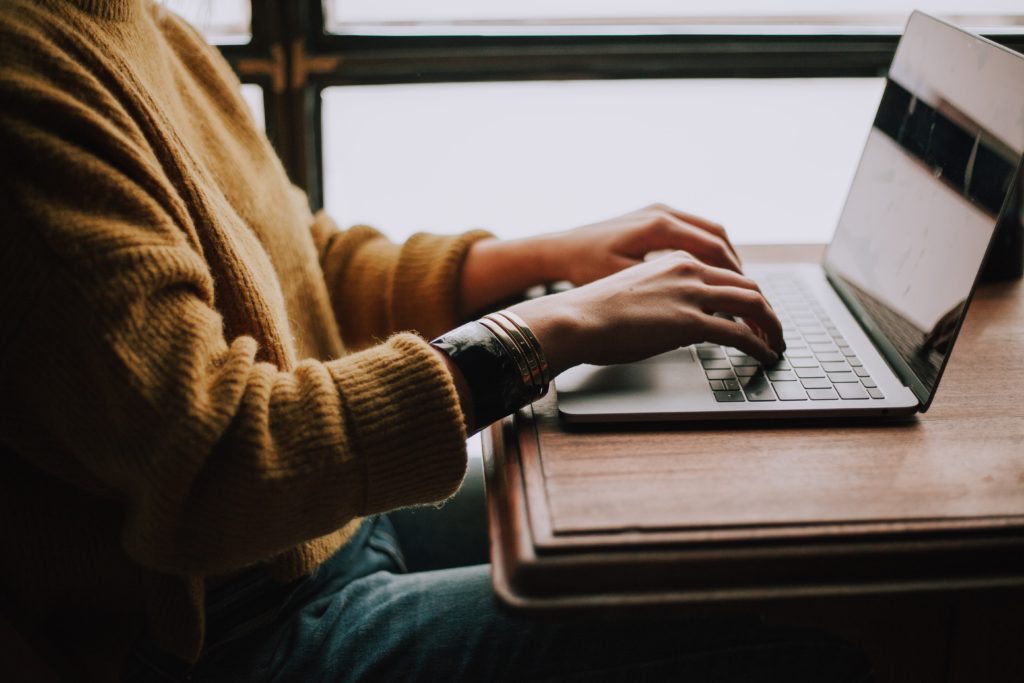 image of a person from the side in a mustard yellow jumper working at a wooden desk on a silver laptop