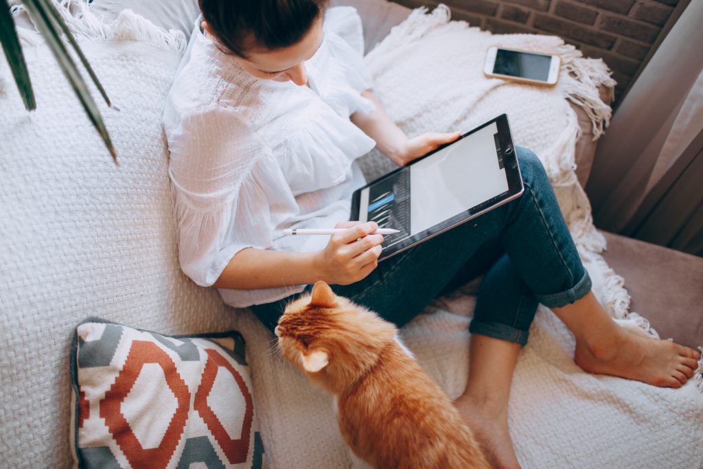 Image from above of a woman sat on a white sofa working on a tablet with a ginger cat next to her